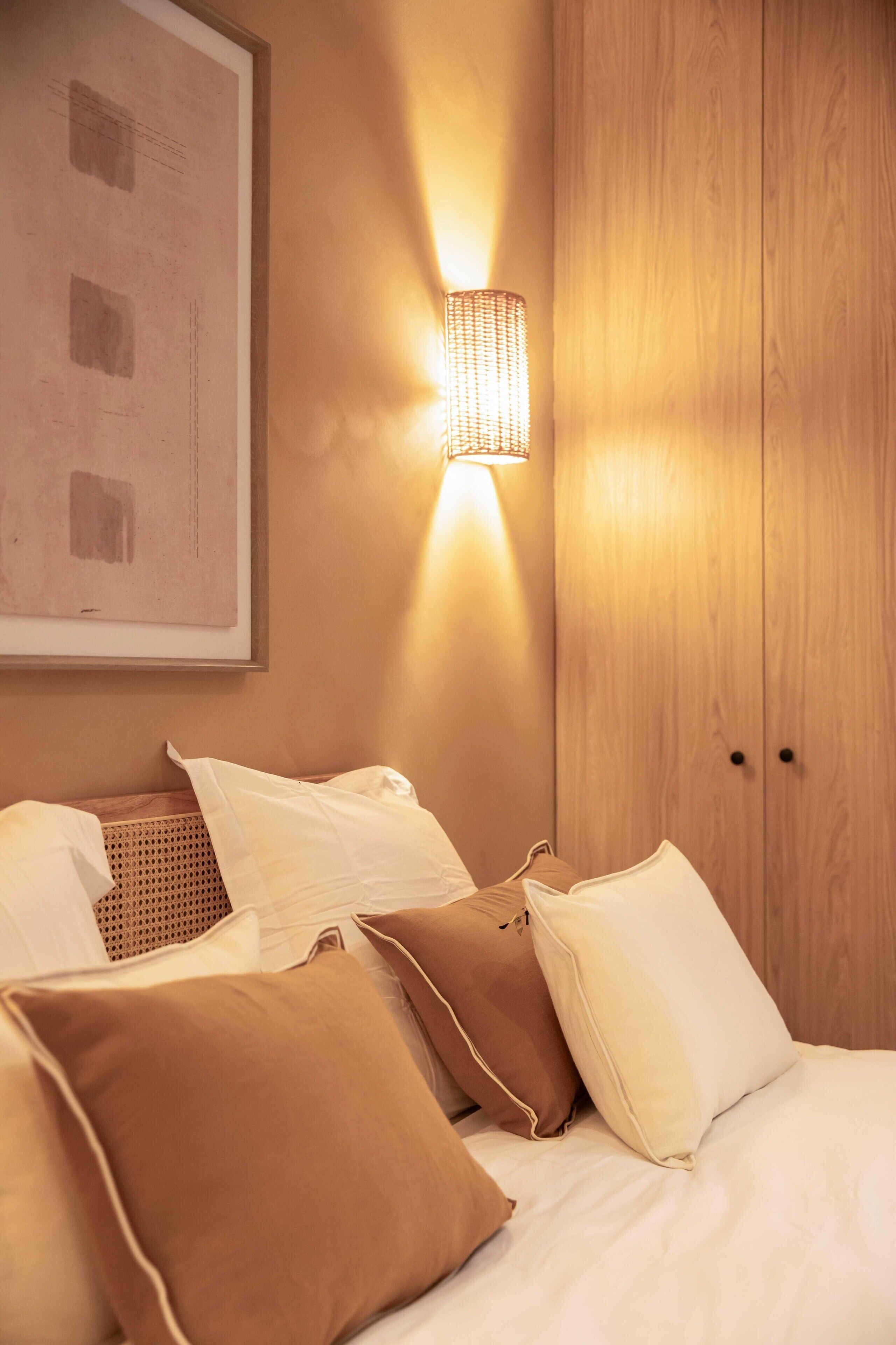 A neatly made bed with white and brown pillows, a wall-mounted light fixture, and a framed picture on the wall.