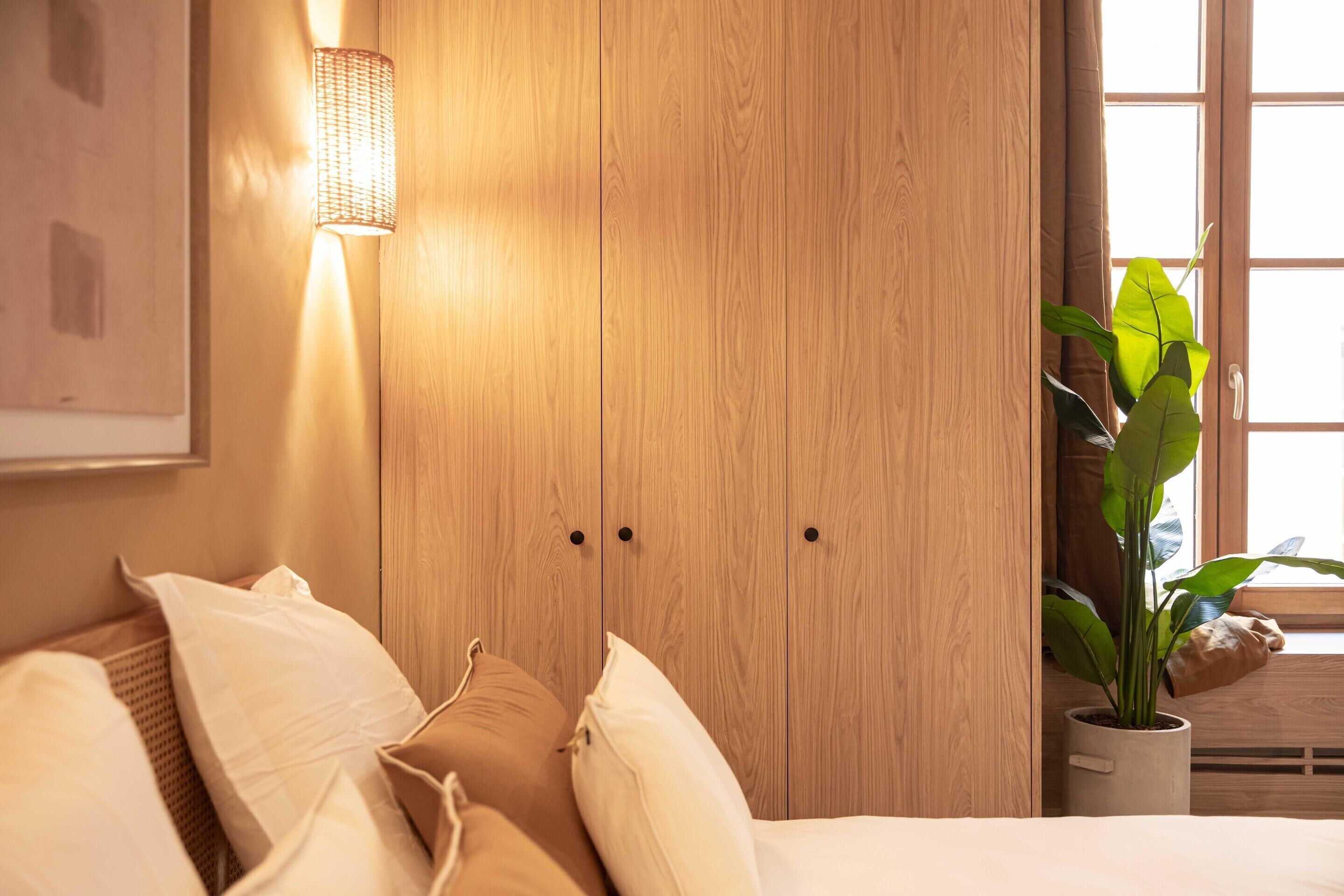 A bedroom with a wooden wardrobe, a bedside lamp, a bed with pillows, and a potted plant by the window.