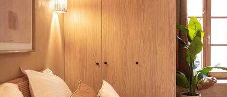 A bedroom with a wooden wardrobe, a bedside lamp, a bed with pillows, and a potted plant by the window.