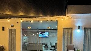 An outdoor seating area with lounge chairs and a table, a glass door entrance, and a television screen visible inside.