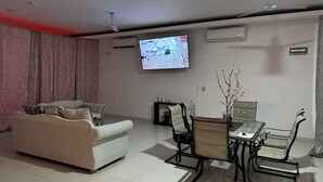 A living room with a sofa, a dining table with chairs, a TV mounted on the wall, and an air conditioning unit.