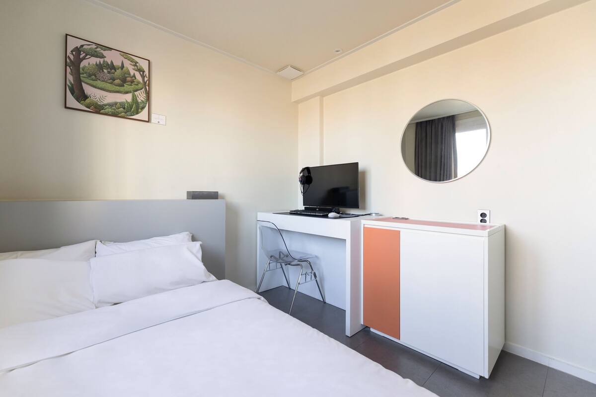 Una habitación de hotel con una cama, un escritorio con una computadora, una silla, un espejo redondo y un cuadro colgado en la pared.