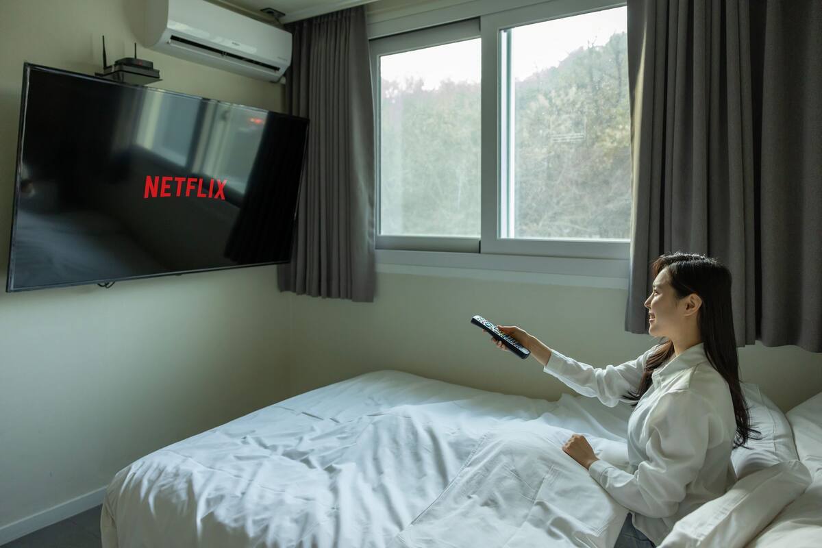 Una habitación de hotel con una cama, un televisor fijado a la pared mostrando 'NETFLIX' y una ventana con cortinas.
