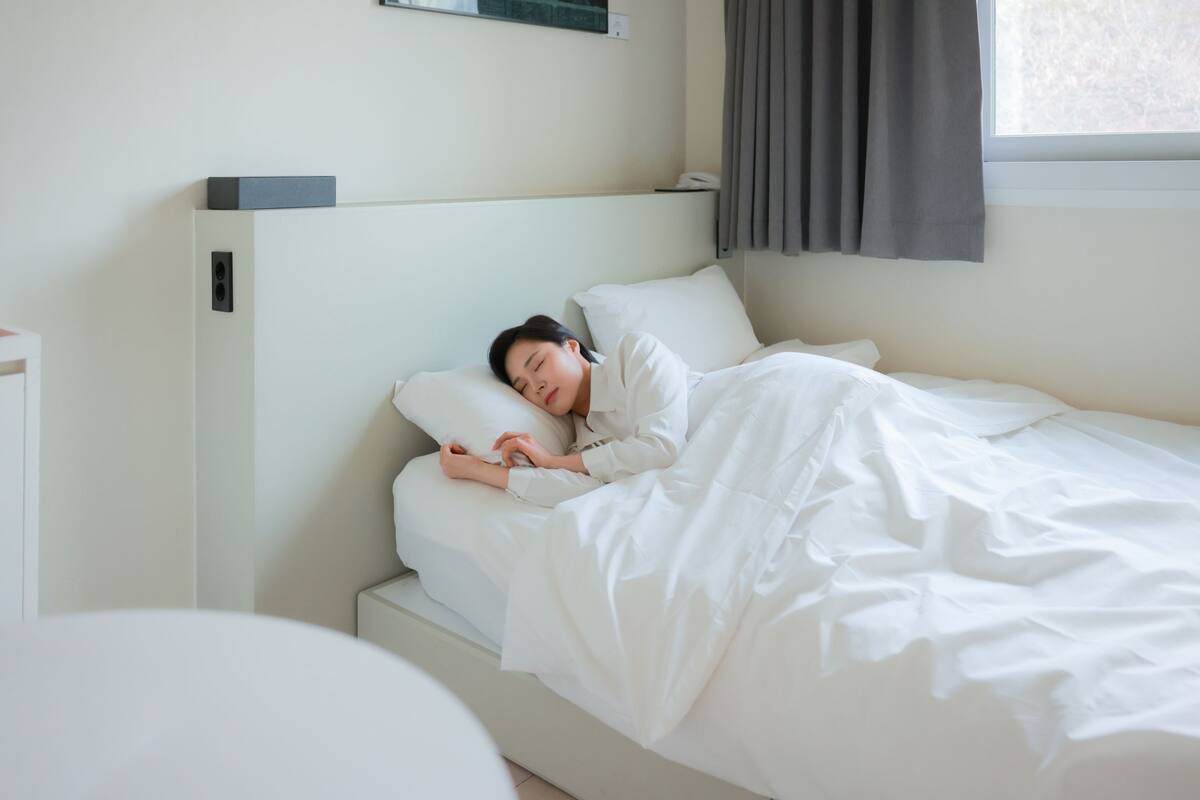 Una cama bien hecha con sábanas blancas en una habitación con cabecera blanca y una ventana con cortinas.