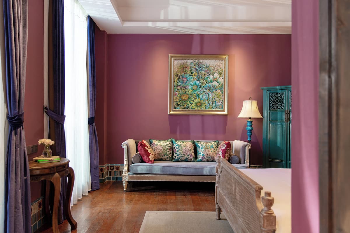 A room with a purple wall, a floral painting, a blue sofa with patterned cushions, a wooden side table, and a teal cabinet.