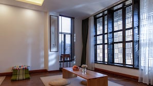 Una habitación con una mesa de madera, esteras tejidas, un taburete pequeño y un ventanal con contraventanas decoradas.