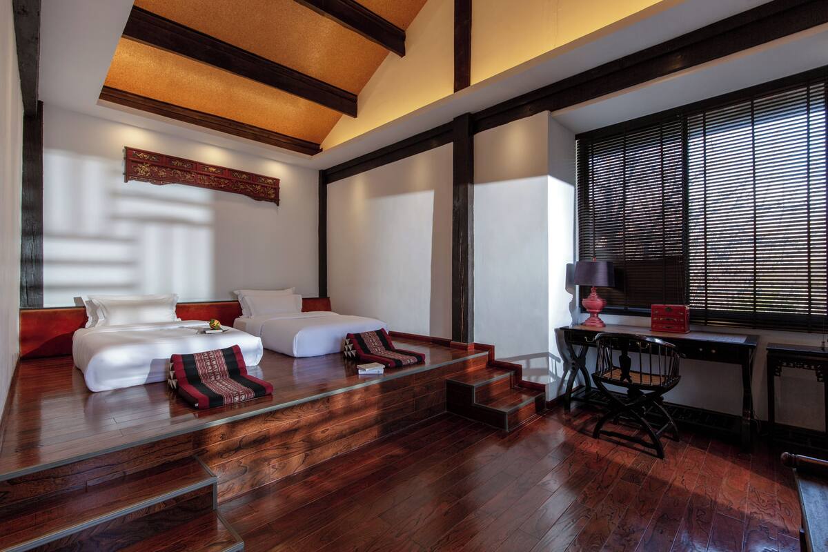A modern hotel room with a wooden floor, a bed with white linens, a desk with a lamp, and a window with blinds.