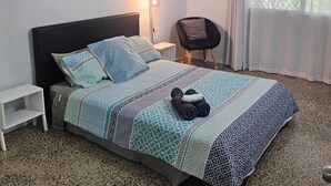 A bedroom with a bed, a chair, a bedside table, a ceiling fan, an air conditioning unit, a window with curtains, and a wall-mounted picture.