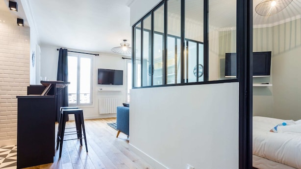 A modern hotel room with a large window, a bed, a desk with a chair, and a television. - Serene Apartment - 1br/4p - Place de la Bastille (Paris)