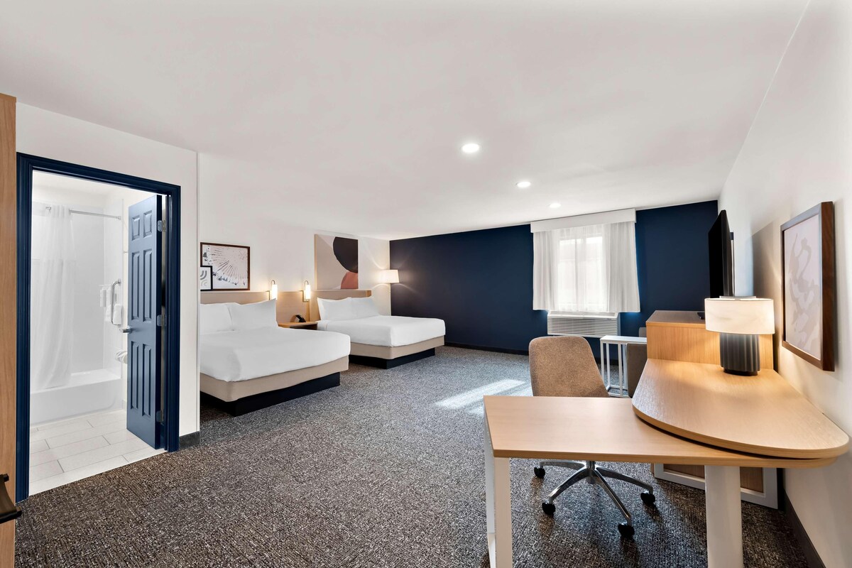 A hotel room with a bed, a desk, a chair, a lamp, and a window.