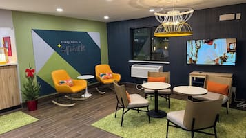 Um lobby de hotel moderno com design geométrico verde e azul na parede, uma grande tela de TV e diversas opções de assento, incluindo cadeiras amarelas e uma mesa redonda.