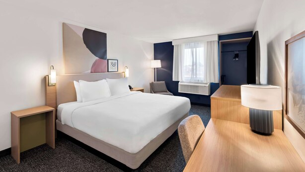 A hotel room with a large bed, bedside tables, a desk, a chair, a lamp, a window with curtains, and a wall-mounted air conditioning unit. - Spark by Hilton Norfolk (Norfolk)