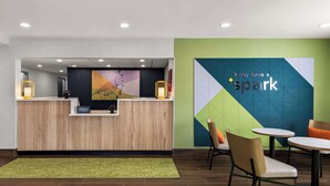A hotel reception area with a wooden reception desk, a digital screen, and a green wall with a geometric design and the text "It only takes a spark". There are two chairs and a small table in the foreground. - Spark by Hilton Norfolk (Norfolk)