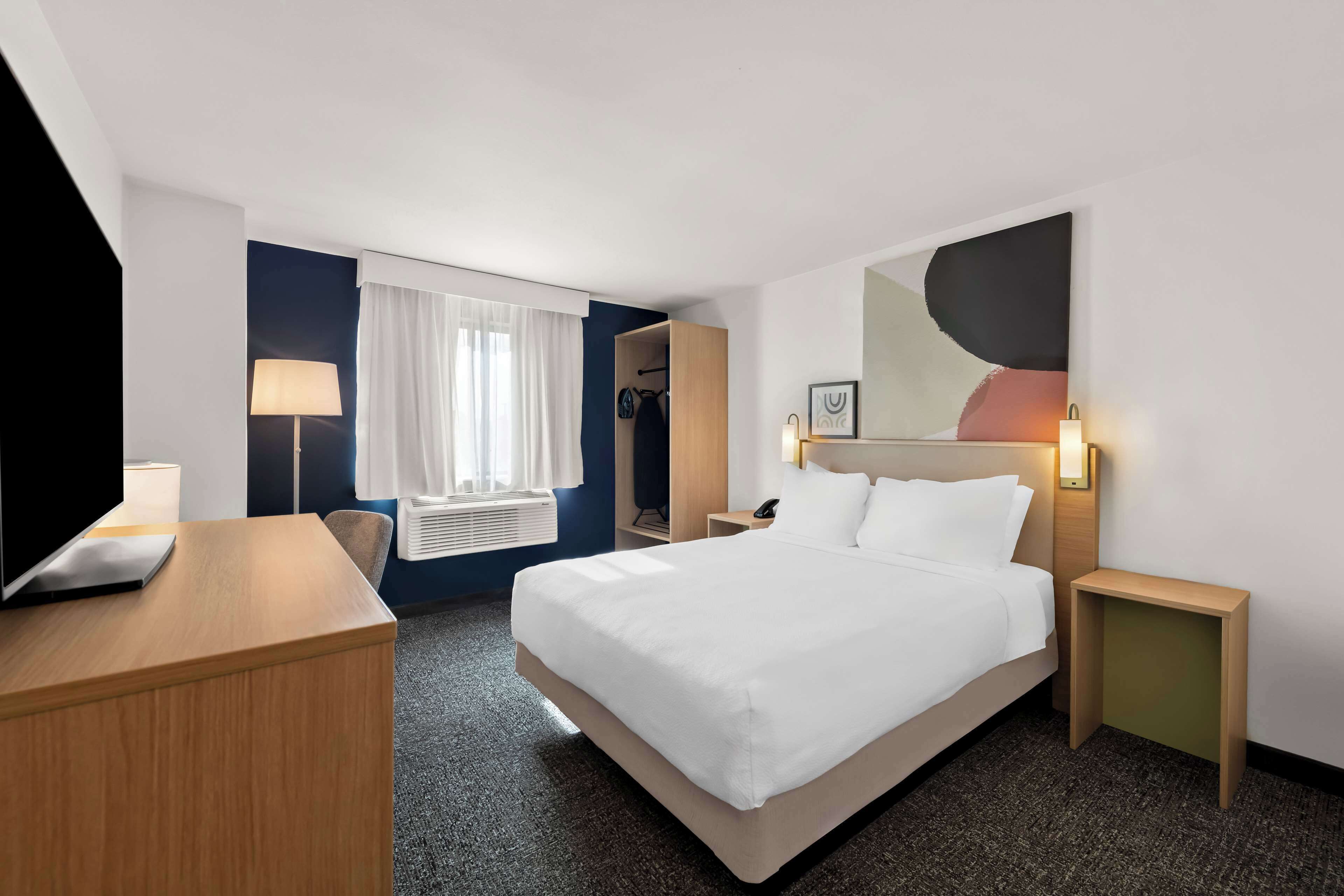 Une chambre d’hôtel comprenant un lit, des tables de chevet, un bureau, une chaise, une lampe, une armoire et une fenêtre avec des rideaux.