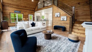 A spacious living area with a white sofa, dark armchairs, and a coffee table. A staircase with a wooden railing leads to an upper level. - Stunning Home Near Nolin Lake: Hot Tub + Fire Pit! (Cub Run)