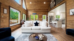 A spacious living room with a white sofa, a coffee table with books and a vase, wooden walls, and large windows offering a view of the outdoors. - Stunning Home Near Nolin Lake: Hot Tub + Fire Pit! (Cub Run)
