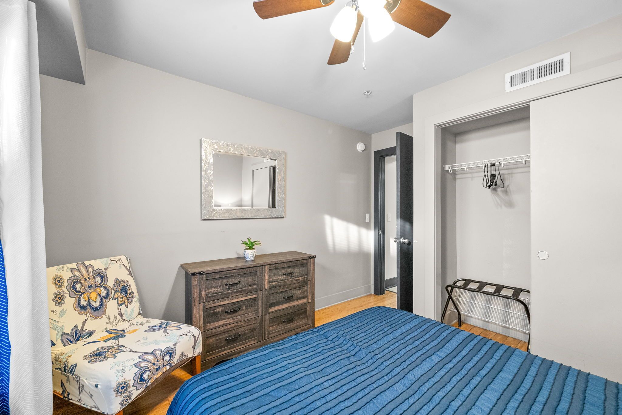 Une chambre à coucher comprenant un lit, une commode, une chaise, un miroir et un ventilateur de plafond.