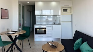 A compact kitchen with white cabinets, a microwave, and a refrigerator. There is a dining area with a table and chairs, and a black sofa with green cushions. - Discover Costa Brava! Rent this cozy studio in Roses with a pool, close to the beach. Book now! (Roses)