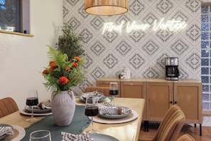 A dining area with a table set for two, a vase with flowers, and a neon sign that reads 'Find Your Vortex'.