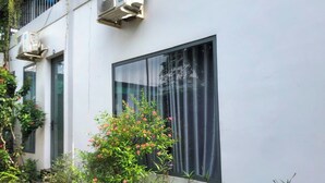 A building with two air conditioning units, a large window with a curtain, and a variety of plants in the foreground. - Homestay Lagi (Phuoc Hoi)
