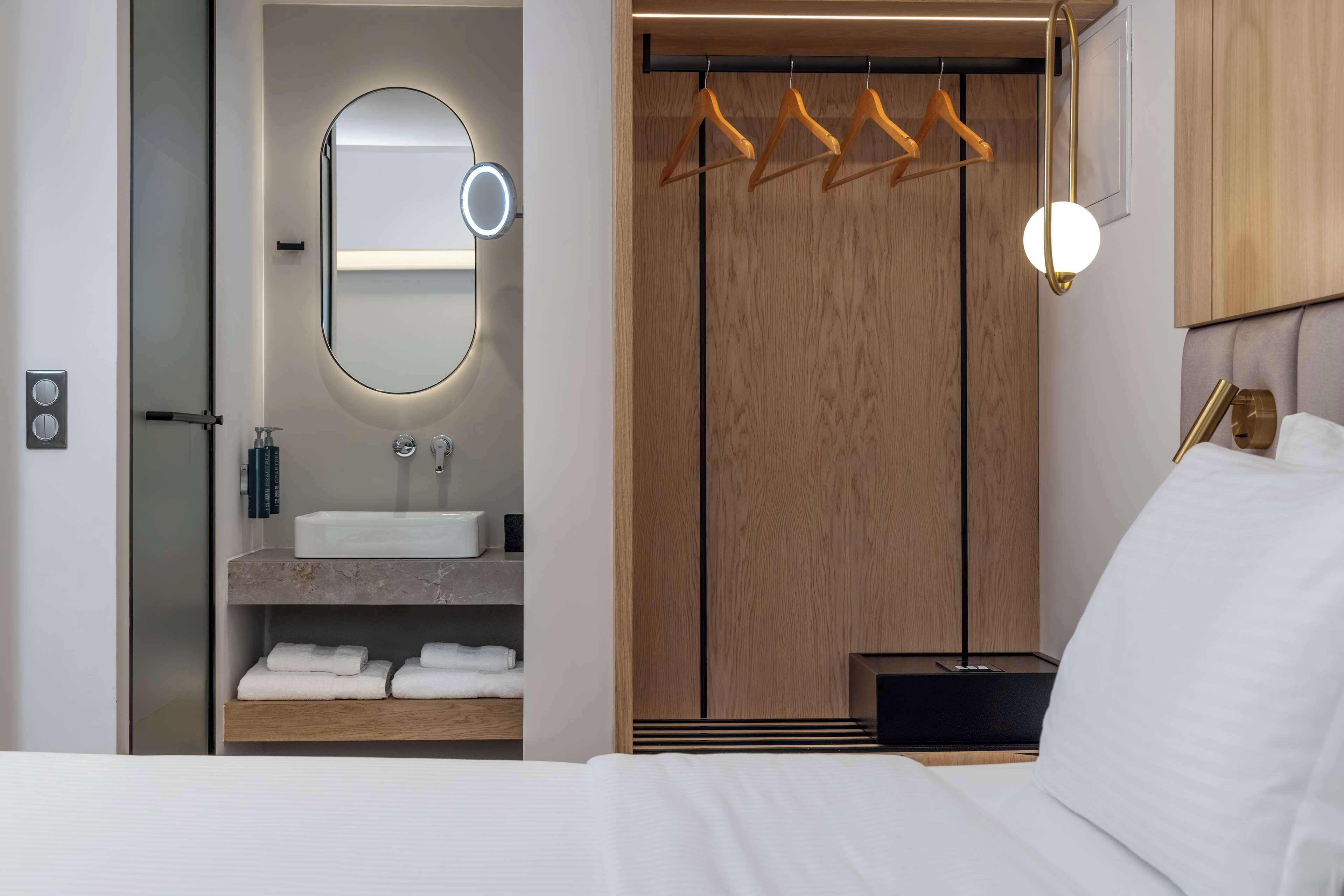 A modern hotel room with a large bed, a wooden wardrobe, a vanity with a mirror, and a hanging light fixture.