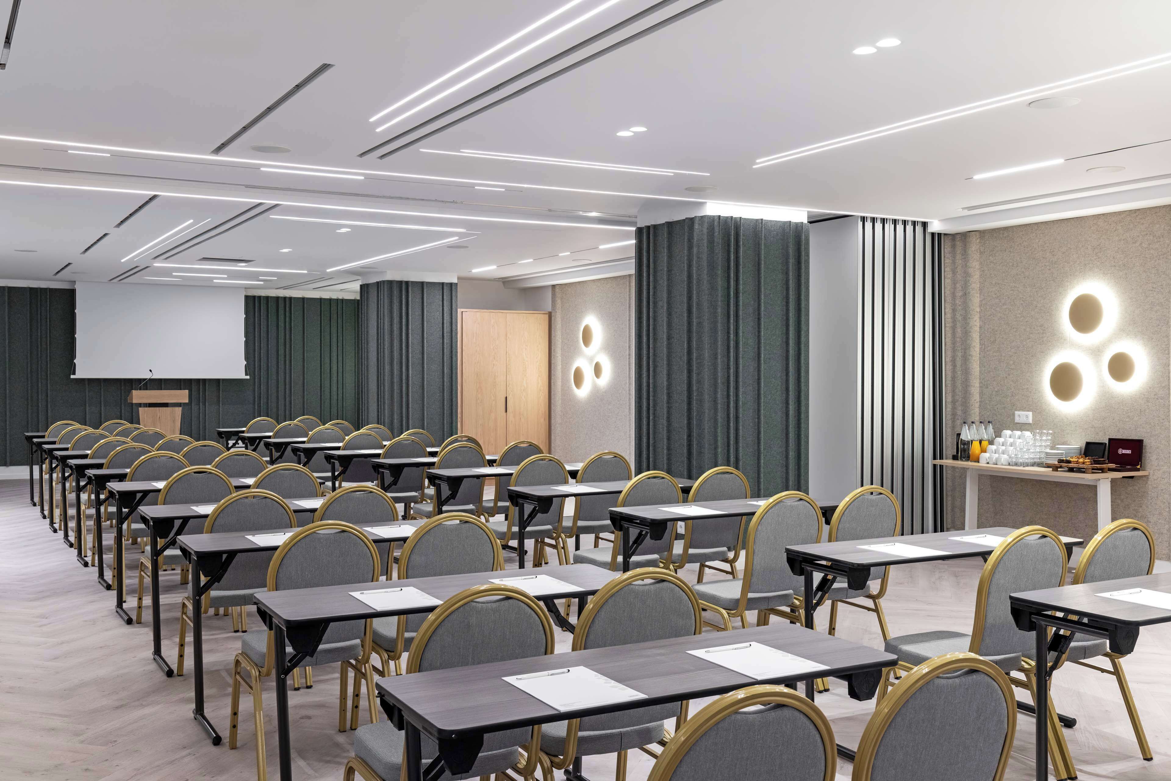 Uma sala de conferências com fileiras de mesas e cadeiras, um púlpito e uma tela de projeção.