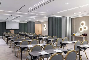 Uma sala de conferências com fileiras de mesas e cadeiras, um púlpito e uma tela de projeção.