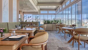 A modern dining area with high ceilings, large windows, and a city view. - Hilton Garden Inn Athens Syggrou Avenue (Kallithea)