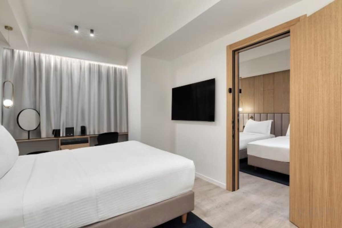 A modern hotel room with a large bed, a desk, a chair, a flat-screen TV, and a wardrobe.