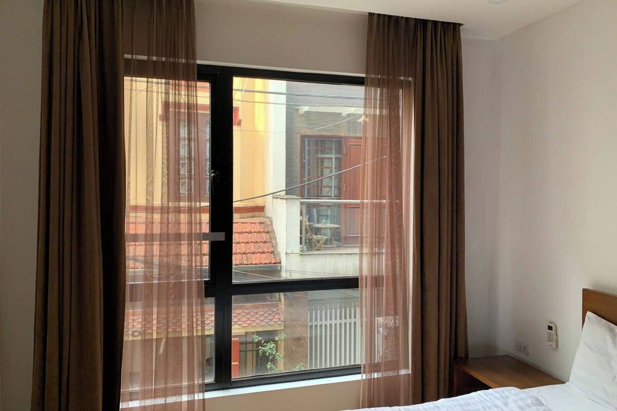 A hotel room with a large window, curtains, a bed, and a bedside table.