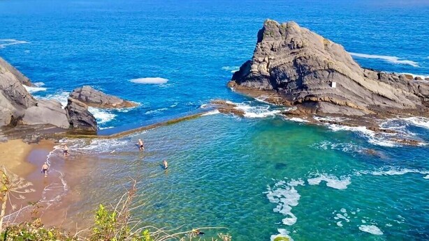 A coastal landscape with clear blue waters, rocky formations, and a few people in the water.