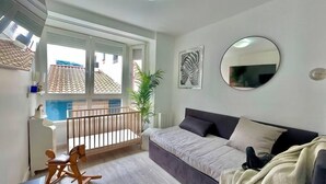 A modern living room with a grey sofa, a wooden rocking chair, a large mirror, and a view of the outside through a balcony.