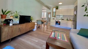 Un salon moderne avec un meuble TV en bois, une table basse en verre et un canapé. On aperçoit une cuisine à l’arrière-plan, avec des meubles blancs et une cuisinière.