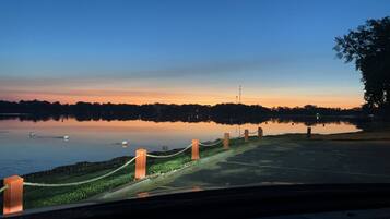Un lago tranquilo al anochecer con cielo despejado, un camino bordeando la orilla y una costa distante con árboles y una línea eléctrica.