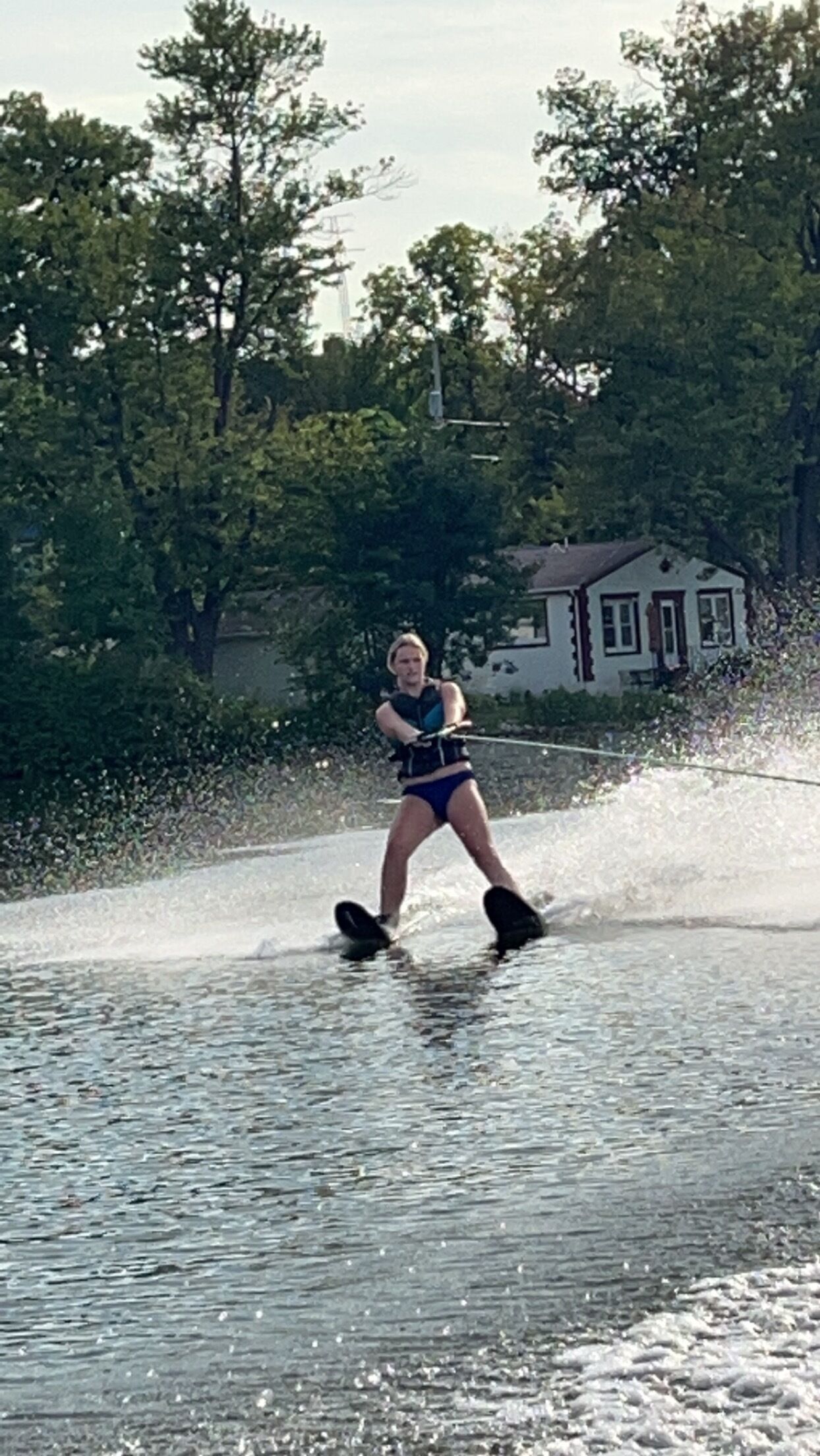 Una persona practicando wakeboard en un cuerpo de agua, con árboles y una casa al fondo.