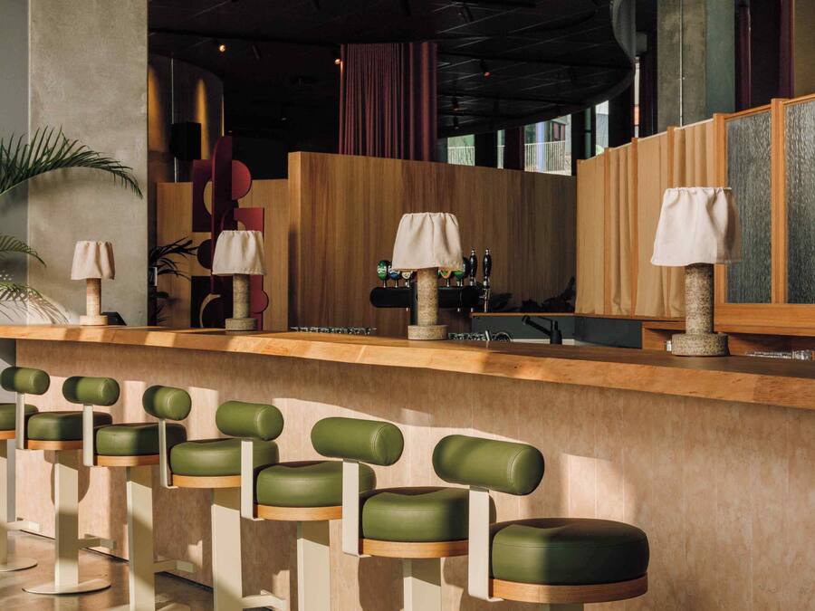 Zona de bar con mostrador de madera, taburetes con cojines verdes y una lámpara decorativa.