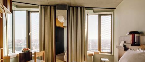 Una habitación de hotel moderna con una cama grande, un escritorio con una silla, una mesita con fruta y vistas a la ciudad a través de amplios ventanales.
