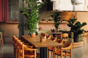 A modern dining area with wooden tables and chairs, potted plants, and a kitchen in the background. - Locke Copenhagen (Copenhagen)
