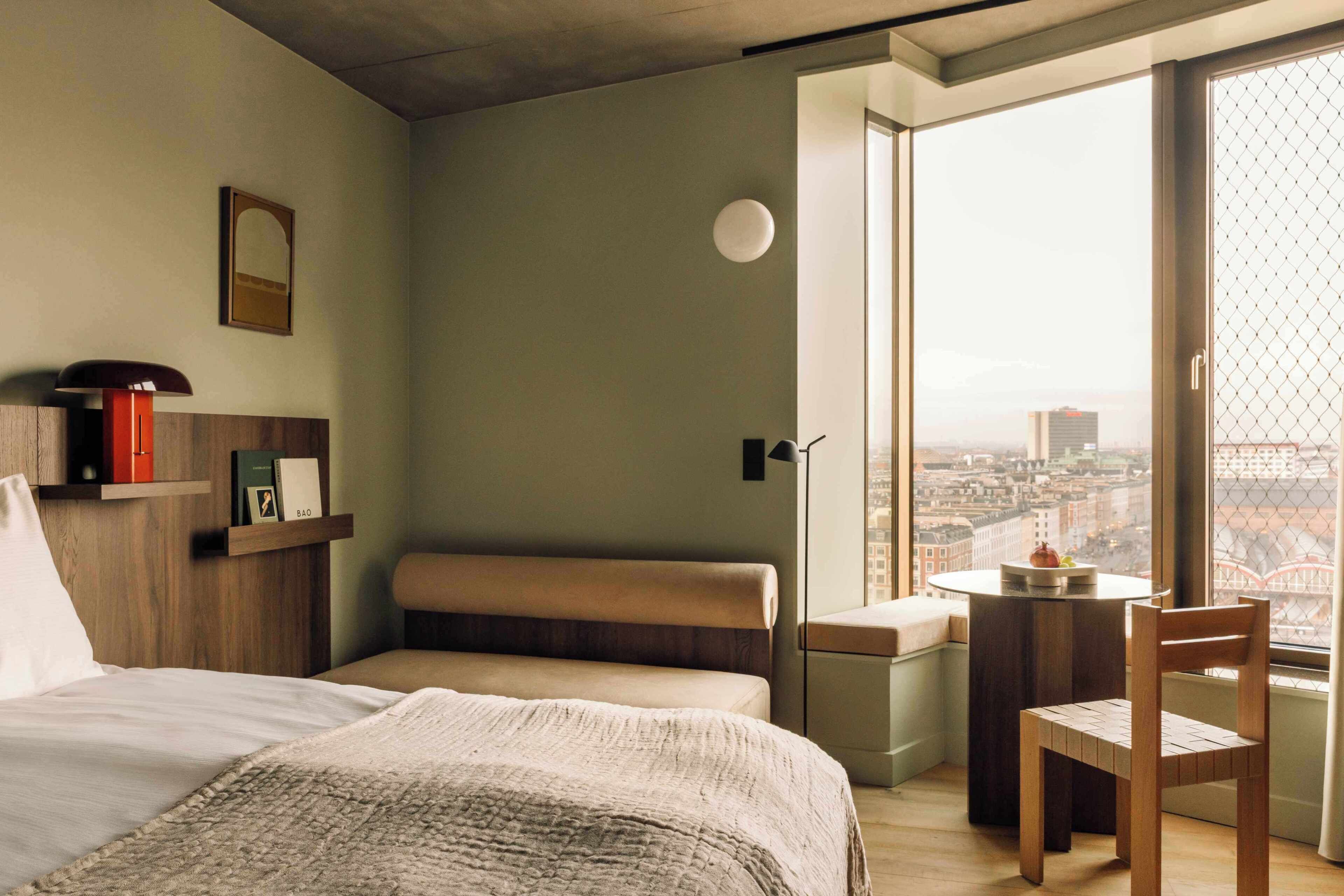 Une chambre d’hôtel moderne dotée d’une grande fenêtre donnant sur la ville, d’un lit confortable, d’une petite table avec une chaise et d’une bibliothèque contenant des livres et un objet rouge.