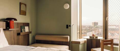 Une chambre d’hôtel moderne dotée d’une grande fenêtre donnant sur la ville, d’un lit confortable, d’une petite table avec une chaise et d’une bibliothèque contenant des livres et un objet rouge.