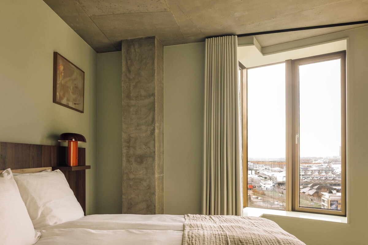 A hotel room with a bed, a nightstand, a lamp, a window with a view of the city, and a curtain.