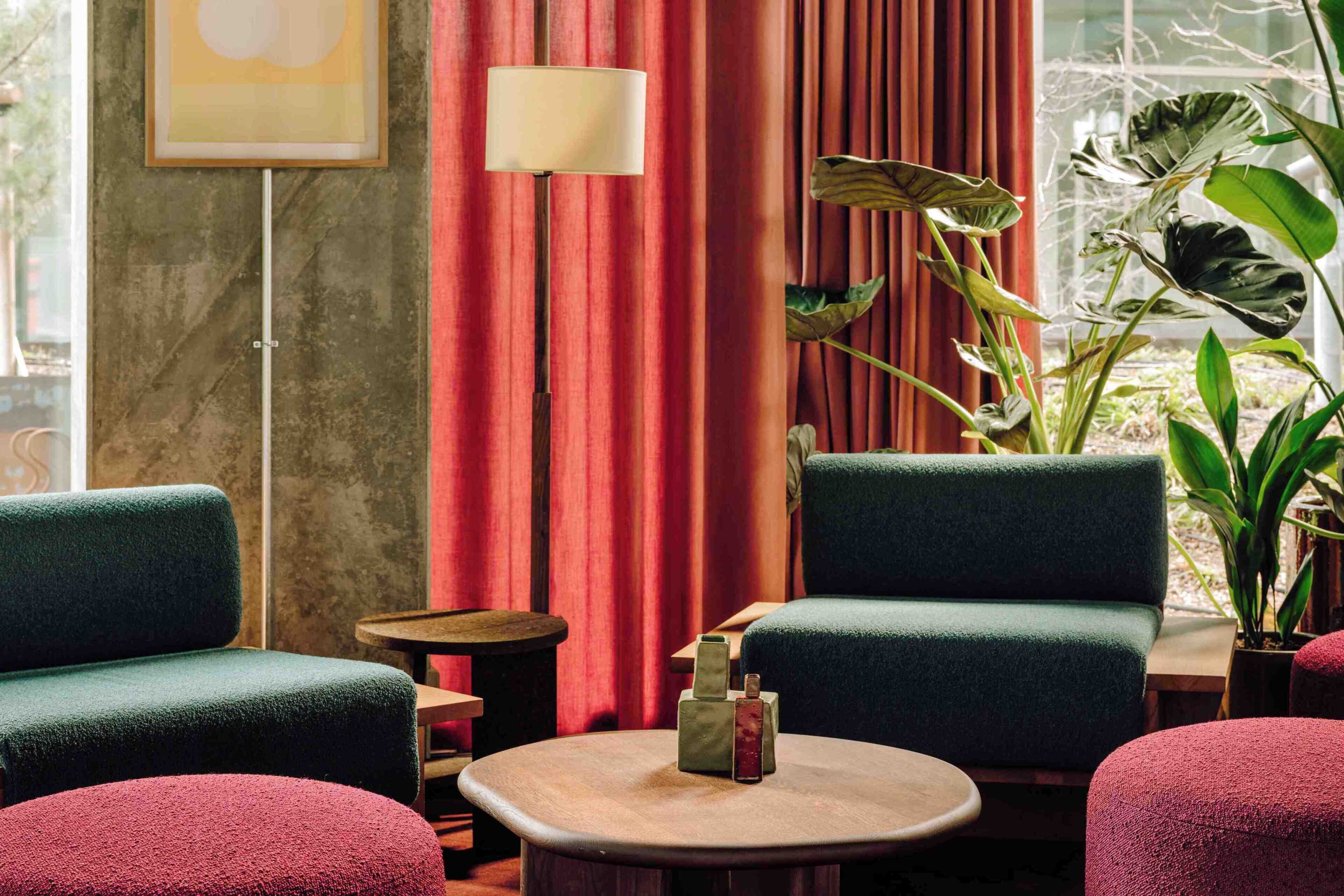 A room with two green armchairs, a round wooden table with a bottle and glasses, a floor lamp, and a large plant.