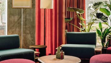 A room with two green armchairs, a round wooden table with a bottle and glasses, a floor lamp, and a large plant.