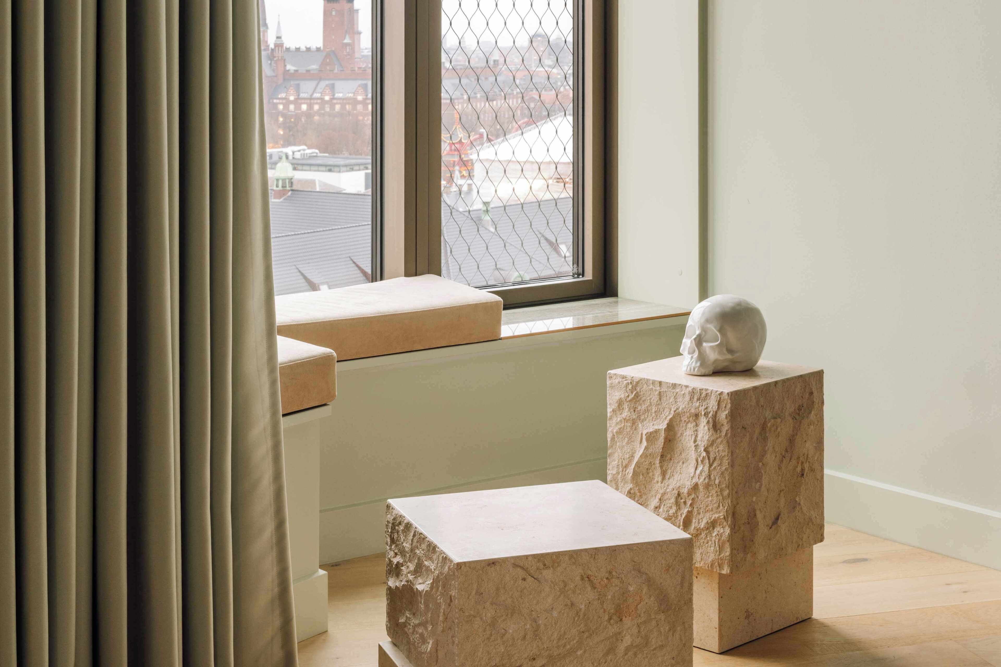 A room with a large window, a white bust sculpture on a pedestal, and a cushioned bench.
