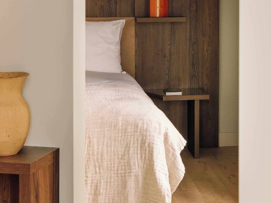 Un dormitorio con cabecera de madera, un jarrón grande sobre una mesita de noche y una mesita con un libro.