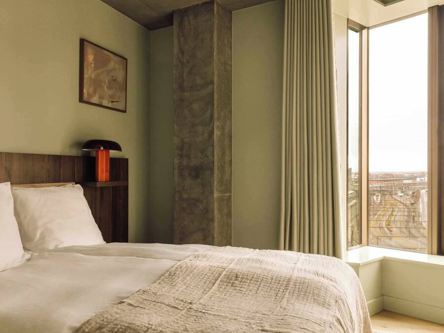 A hotel room with a large bed, a wooden headboard, a nightstand with a lamp, a window with a view of the city, and a framed picture on the wall.