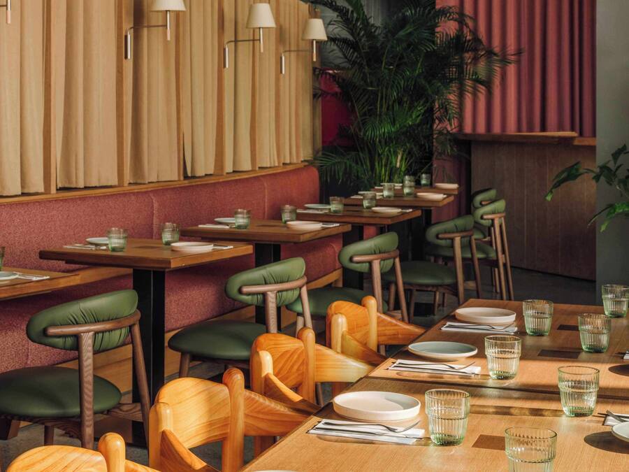 Interior de restaurante con mesas y sillas de madera, asientos acolchados verdes y un telón rojo de fondo.