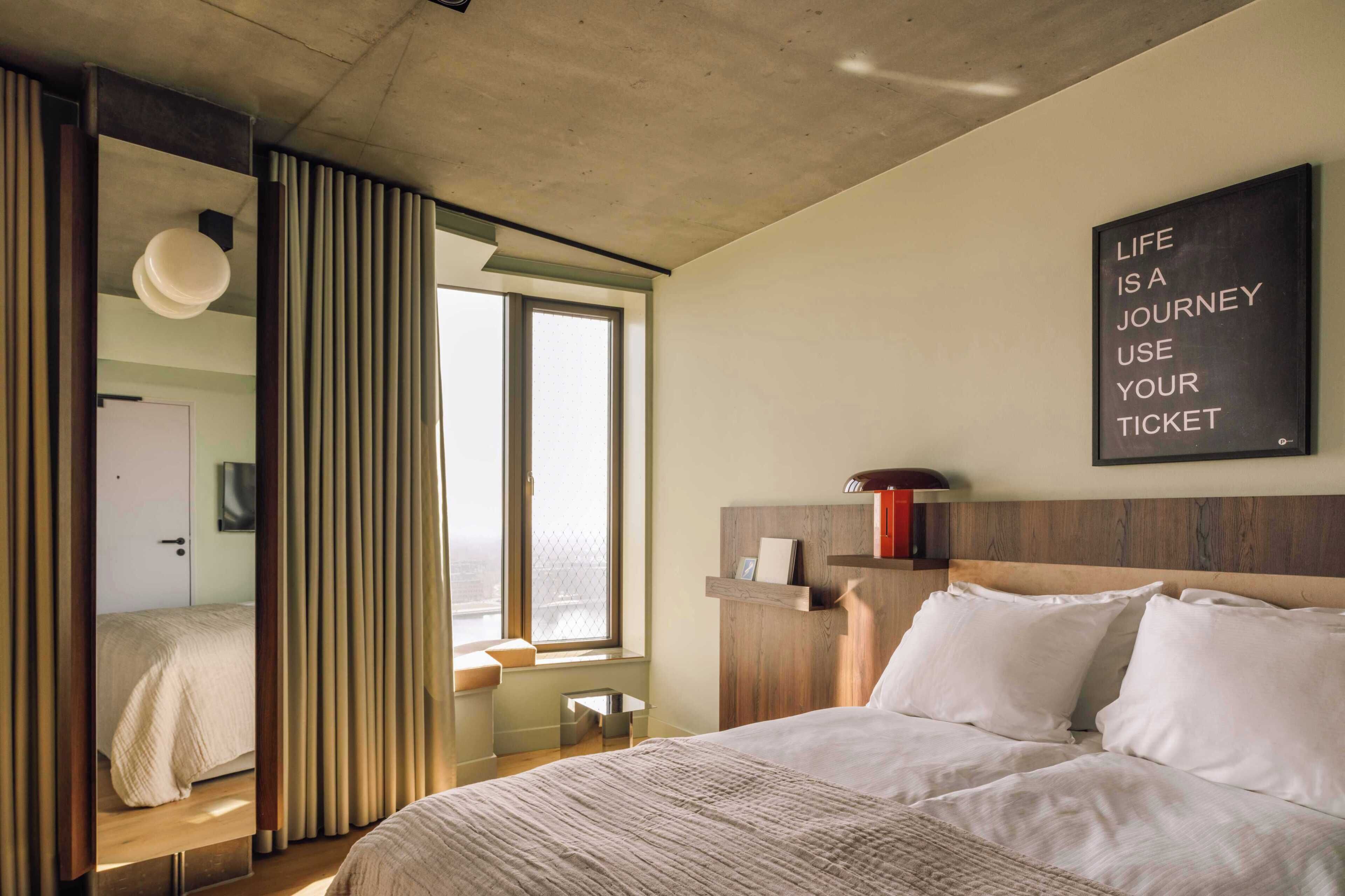A modern hotel room with a large bed, a window with blinds, a wooden headboard, and a motivational quote on the wall.
