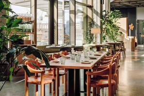 Un comedor moderno con mesas y sillas de madera, ventanas amplias y plantas de interior.