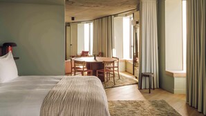 A bedroom with a large bed, a wooden dining table, and a view of the outside through large windows.